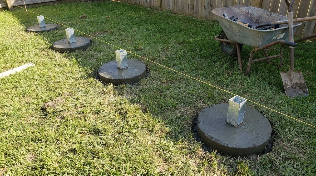 Fresh concrete pergola footings with galvanized raised post bases set in a lawn