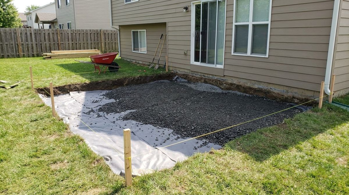 Excavated patio area showing compacted crushed gravel base, string lines and stakes beside a lawn