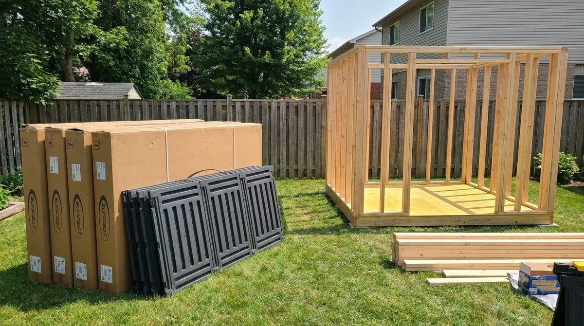 Boxed resin shed kit next to a half-framed wood shed on a backyard floor platform