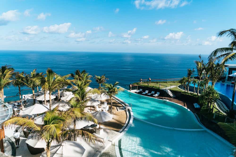 Aerial view of a tropical resort pool surrounded by palm trees next to a turquoise ocean beach in Southeast Asia