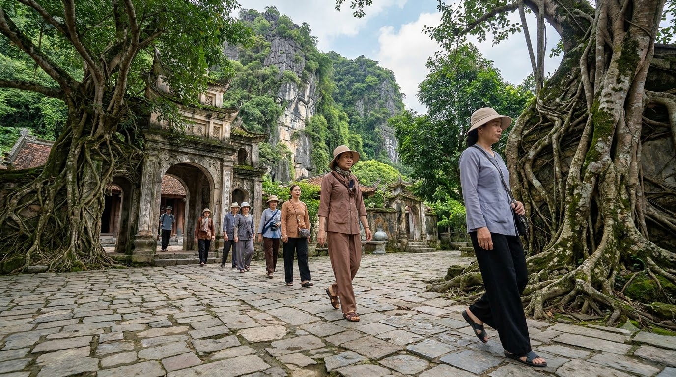 Visitors in respectful attire walking through the stone-paved courtyard of Ha Chua Lower Pagoda with ancient banyan tree roots and limestone cliff backdrop at Bich Dong