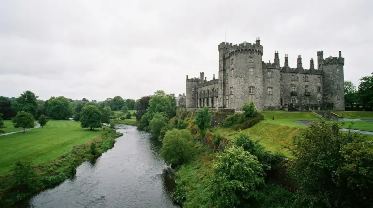 A grey stone castle with round towers above a river and parkland, Kilkenny, Ireland