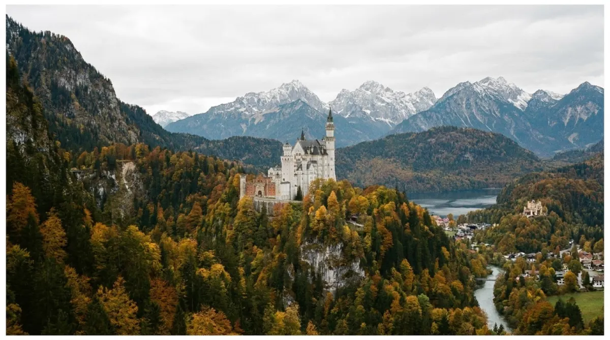 A white fairytale castle with turrets on a wooded hill below alpine mountains, Bavaria, Germany