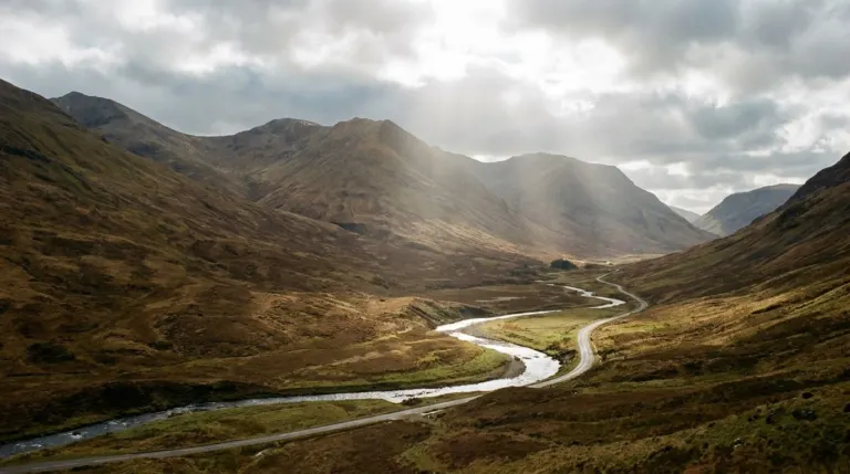 Bare brown mountains above a winding road and a river valley under broken cloud, Scottish Highlands