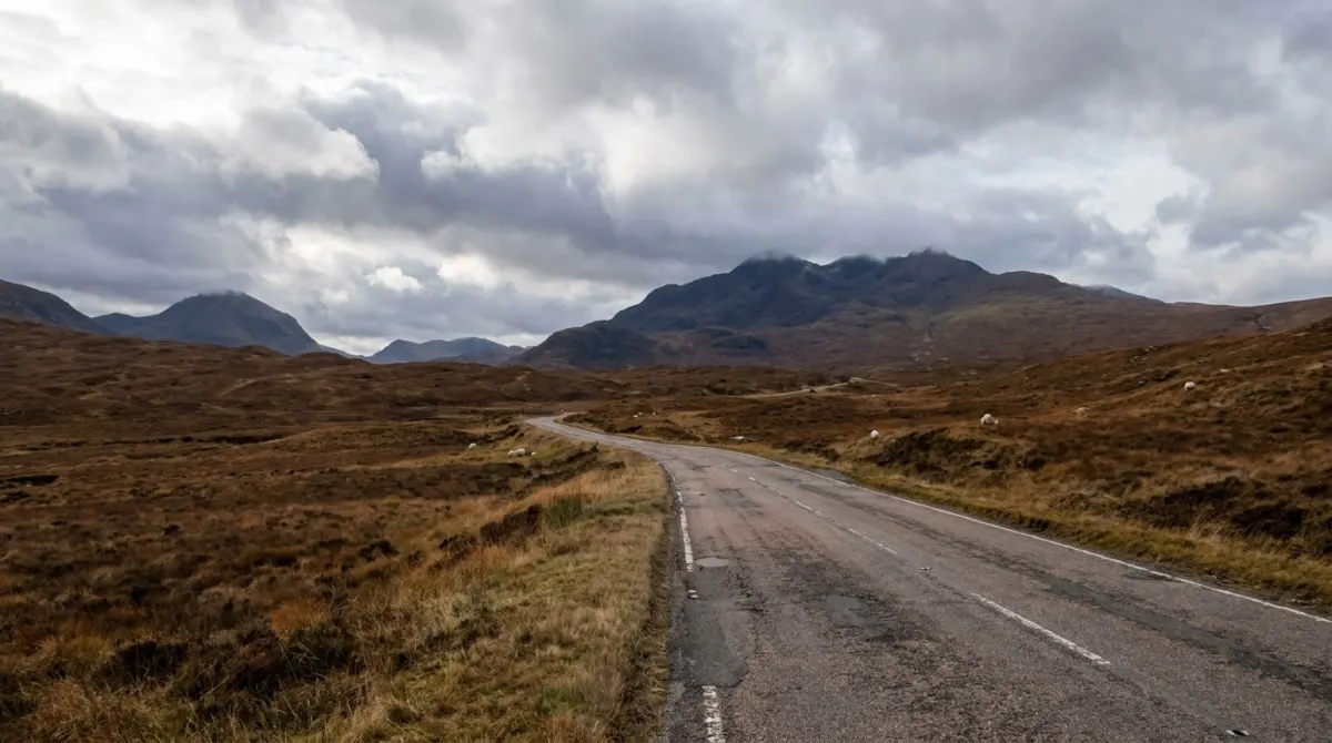 A road curving through open Highland moorland with mountains ahead under broken cloud, Scotland