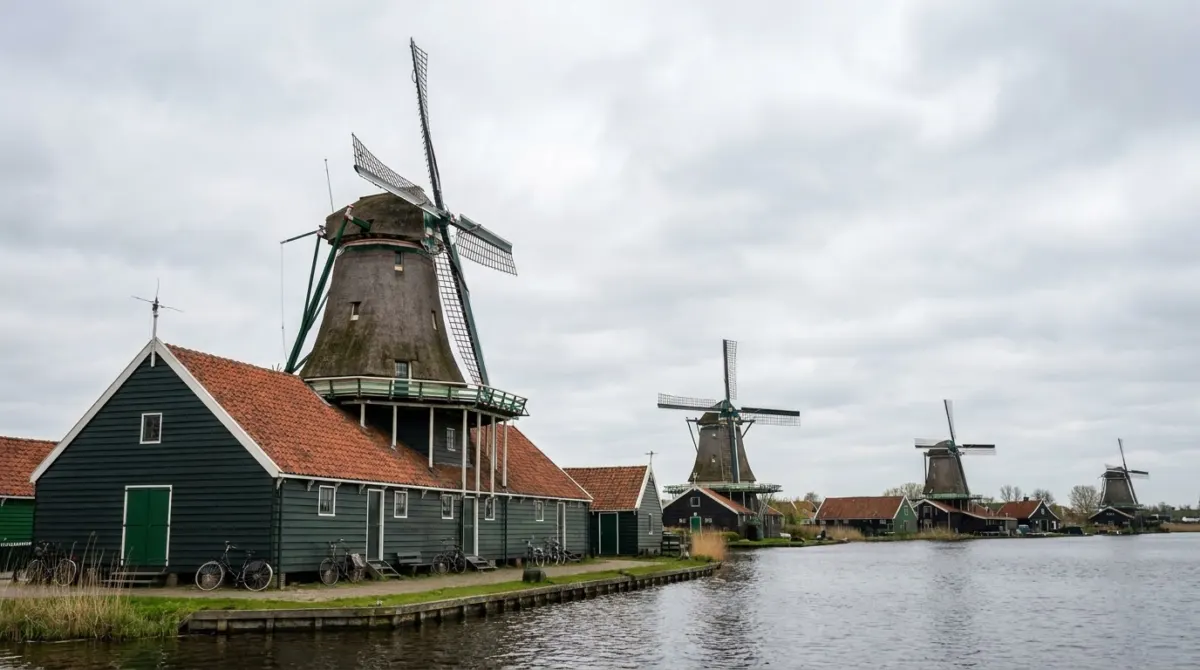 A row of wooden windmills and green houses beside a river under a wide cloudy sky, Zaanse Schans, Netherlands