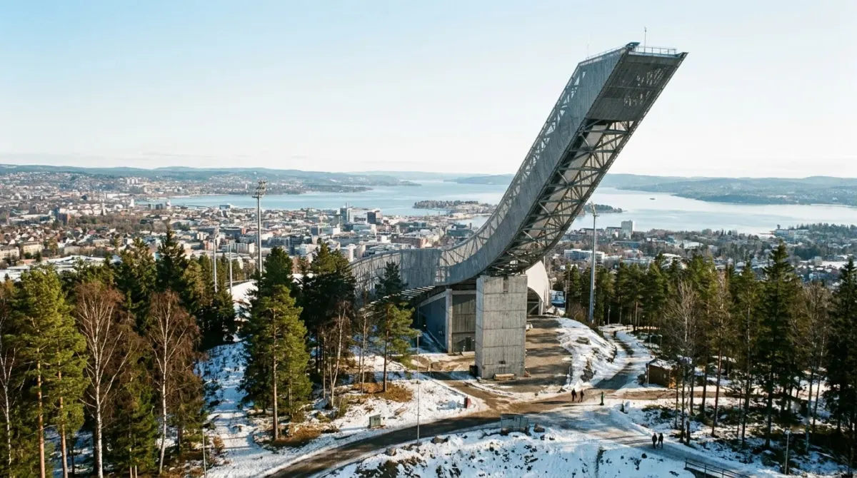 A large modern ski jump structure on a wooded hill above a city and fjord, Holmenkollen, Oslo