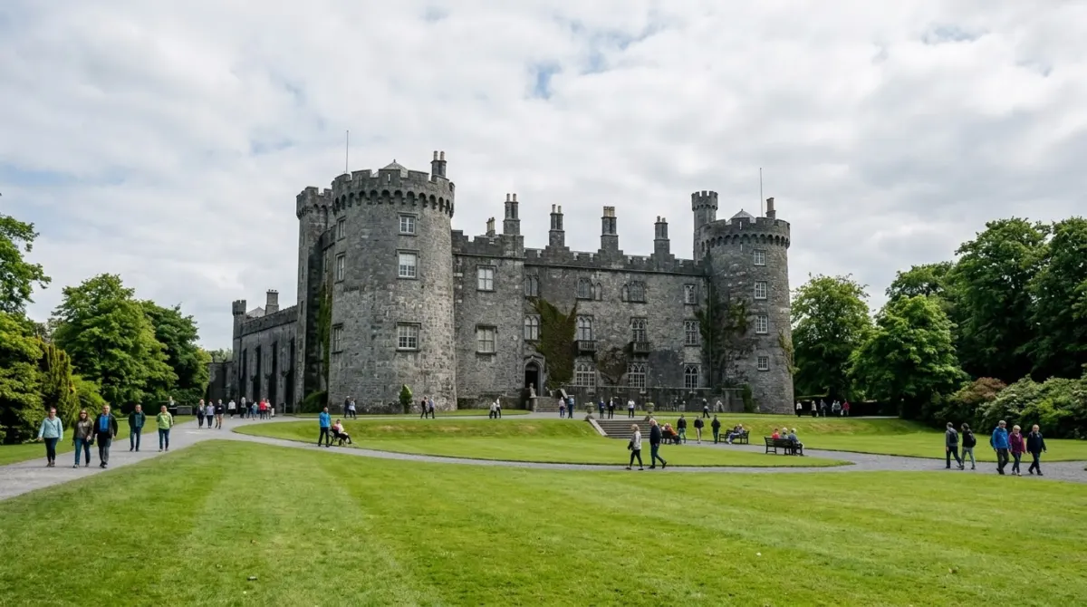 A large grey stone castle with round towers above a wide green lawn and people strolling, Kilkenny Castle, Ireland