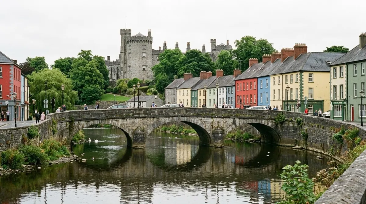A stone bridge over a calm river with a row of colourful townhouses and a castle tower behind, Kilkenny, Ireland