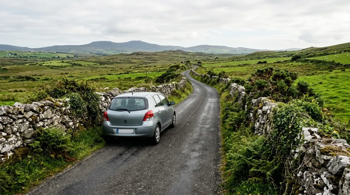A small rental car on the left side of a narrow winding road between stone walls and green fields, west of Ireland