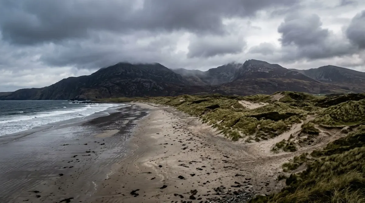 A wild empty beach with dunes and dark mountains behind under a moody sky, County Donegal, Ireland
