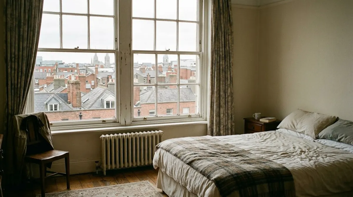 A simple hotel bedroom with a large sash window overlooking Dublin rooftops in soft daylight