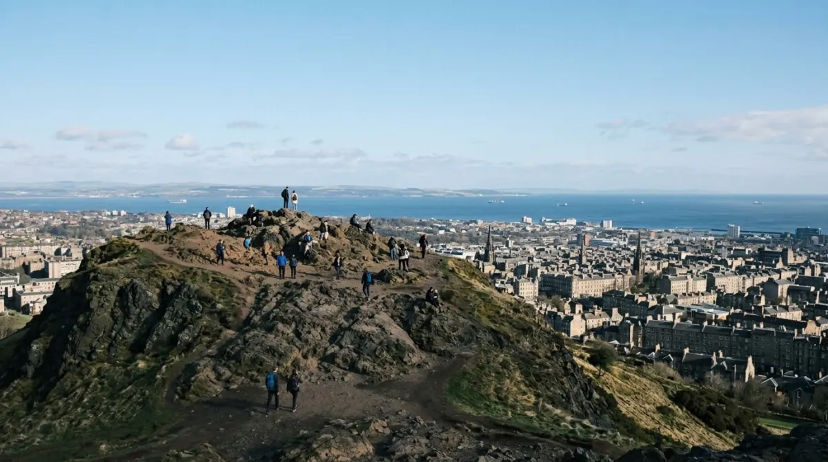 Walkers small on a rocky summit looking over the city rooftops toward the sea on a clear day, Arthur's Seat, Edinburgh, Scotland