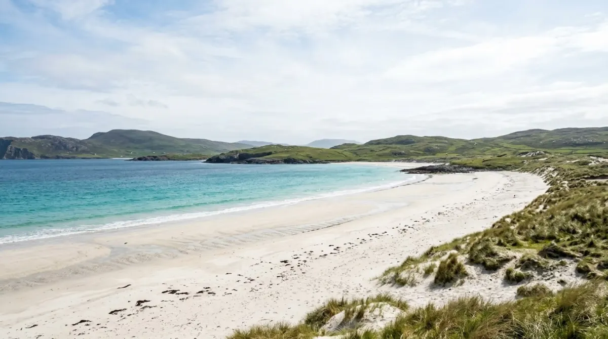 A white sand beach with turquoise water and low green hills under a bright sky, Outer Hebrides, Scotland