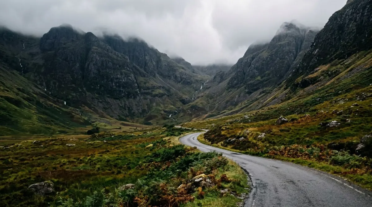 A narrow road winding through a steep green glen under low cloud and dark mountains, Glencoe, Scotland