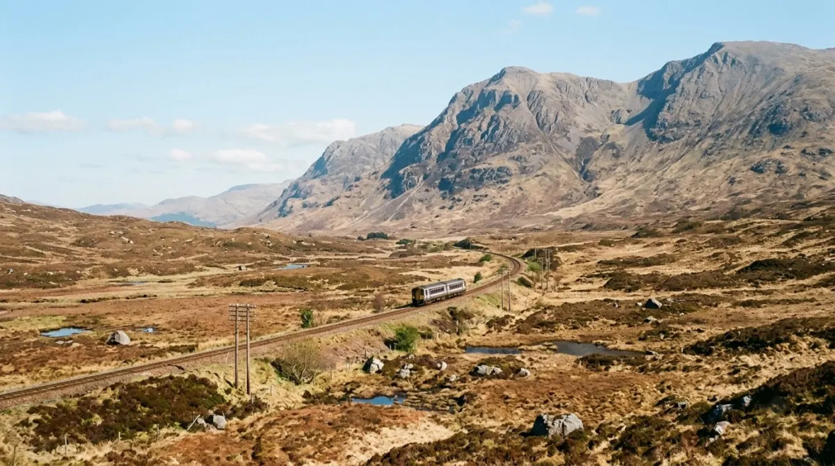 A small train crossing open moorland below bare mountains on a clear day, West Highland Line, Fort William, Scotland