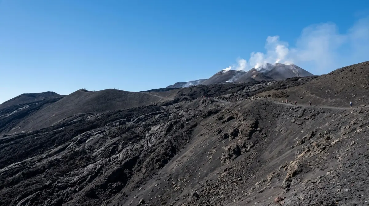 Barren black lava slopes and steaming summit craters under a clear sky with distant walkers on a ridge, Etna, Italy