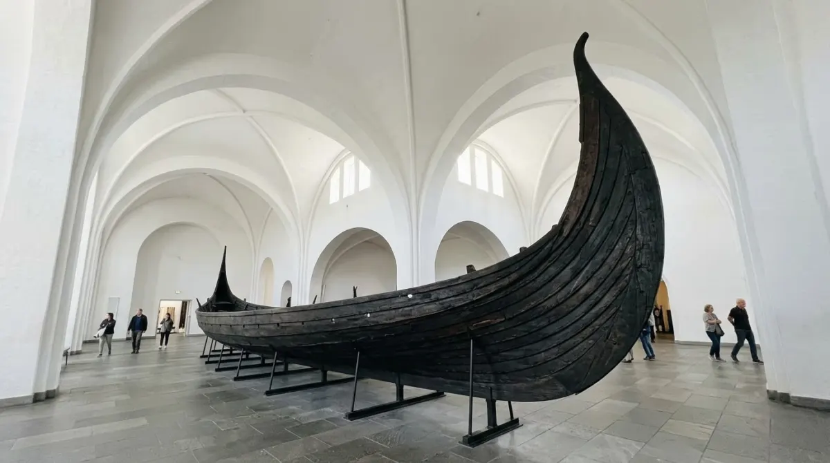 A dark wooden longship with a curved prow displayed in a white vaulted museum hall, Bygdøy, Oslo, Norway