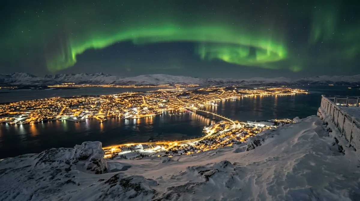 Green aurora arching over the lit city and a bridge seen from a snowy mountain viewpoint, Fjellheisen, Tromsø, Norway