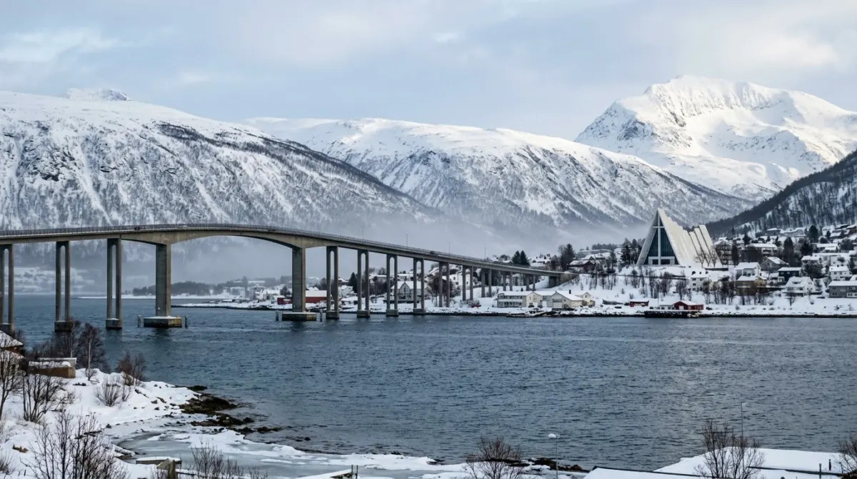 A white triangular church beyond a long bridge across the water with snowy mountains behind, Tromsdalen, Tromsø, Norway