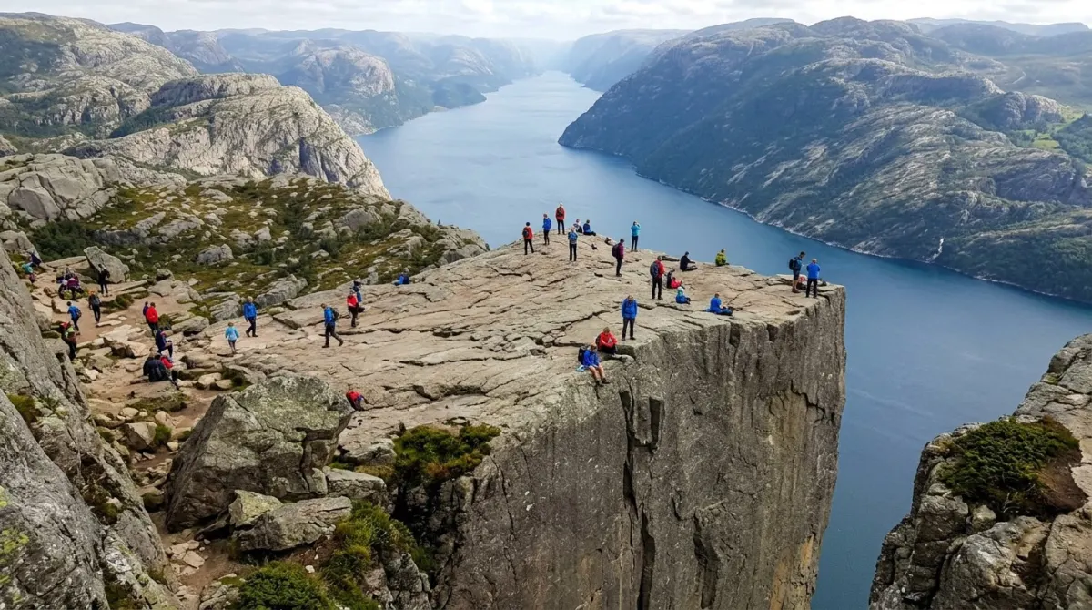 A flat-topped granite cliff platform hanging high above a deep blue fjord with tiny hikers near the edge, Preikestolen, Stavanger, Norway