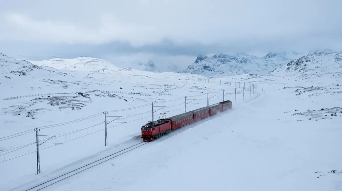 A red train crossing a snow-covered mountain plateau under a pale winter sky on the line between the two cities, Bergen, Norway